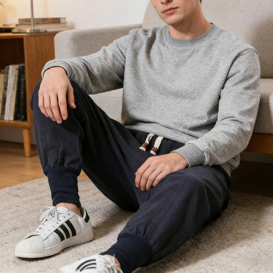 A man sitting on a carpet in a living room setting, wearing a light gray crewneck sweatshirt, dark navy jogger pants