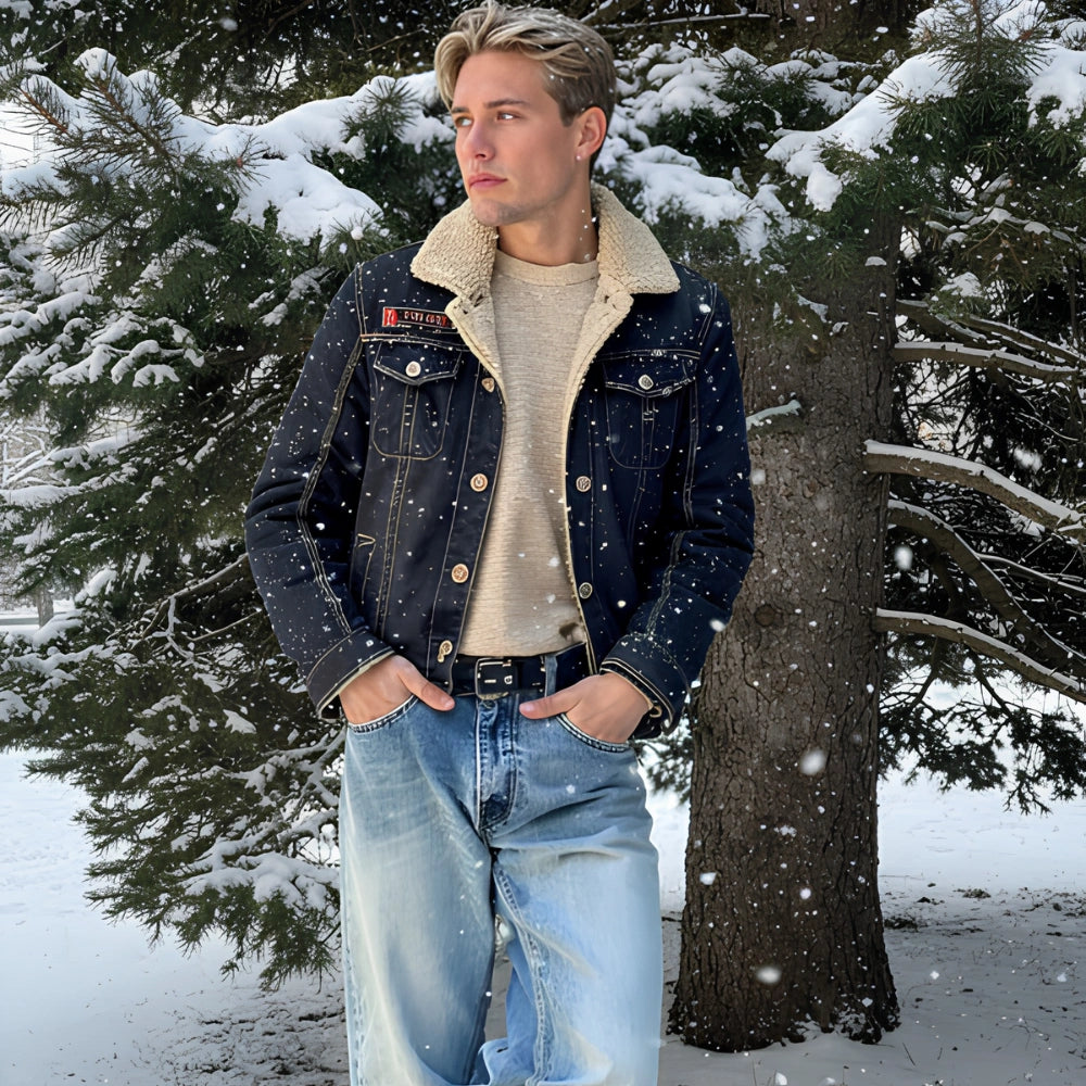 A young man standing outdoors in the snow wearing a dark indigo denim jacket with a cream sherpa collar, a beige knit sweater, and light blue jeans, with snow-covered pine trees in the background.
