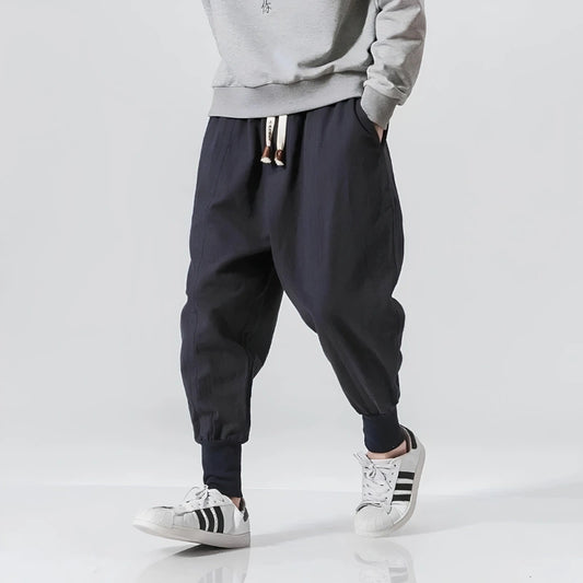 A person wearing loose dark navy jogger-style pants with a drawstring waist, paired with a light gray sweatshirt and white sneakers with black stripes, photographed against a clean light gray studio background.